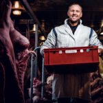 Cheerful butcher in workwear holding a box with meat pieces while standing in the midst of meat carcasses